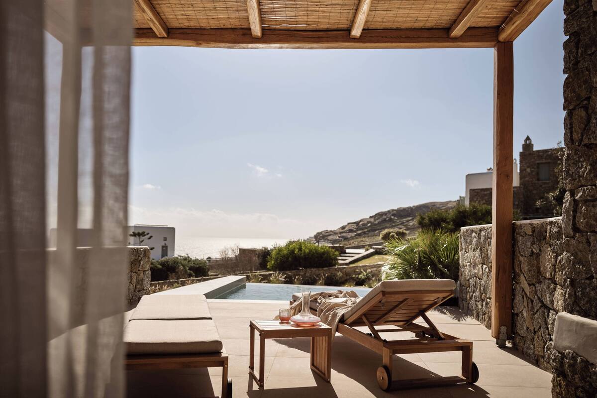 A poolside area with lounge chairs, a table, and a view of the sea and hills.