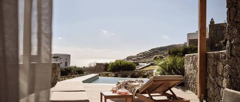 A poolside area with lounge chairs, a table, and a view of the sea and hills.