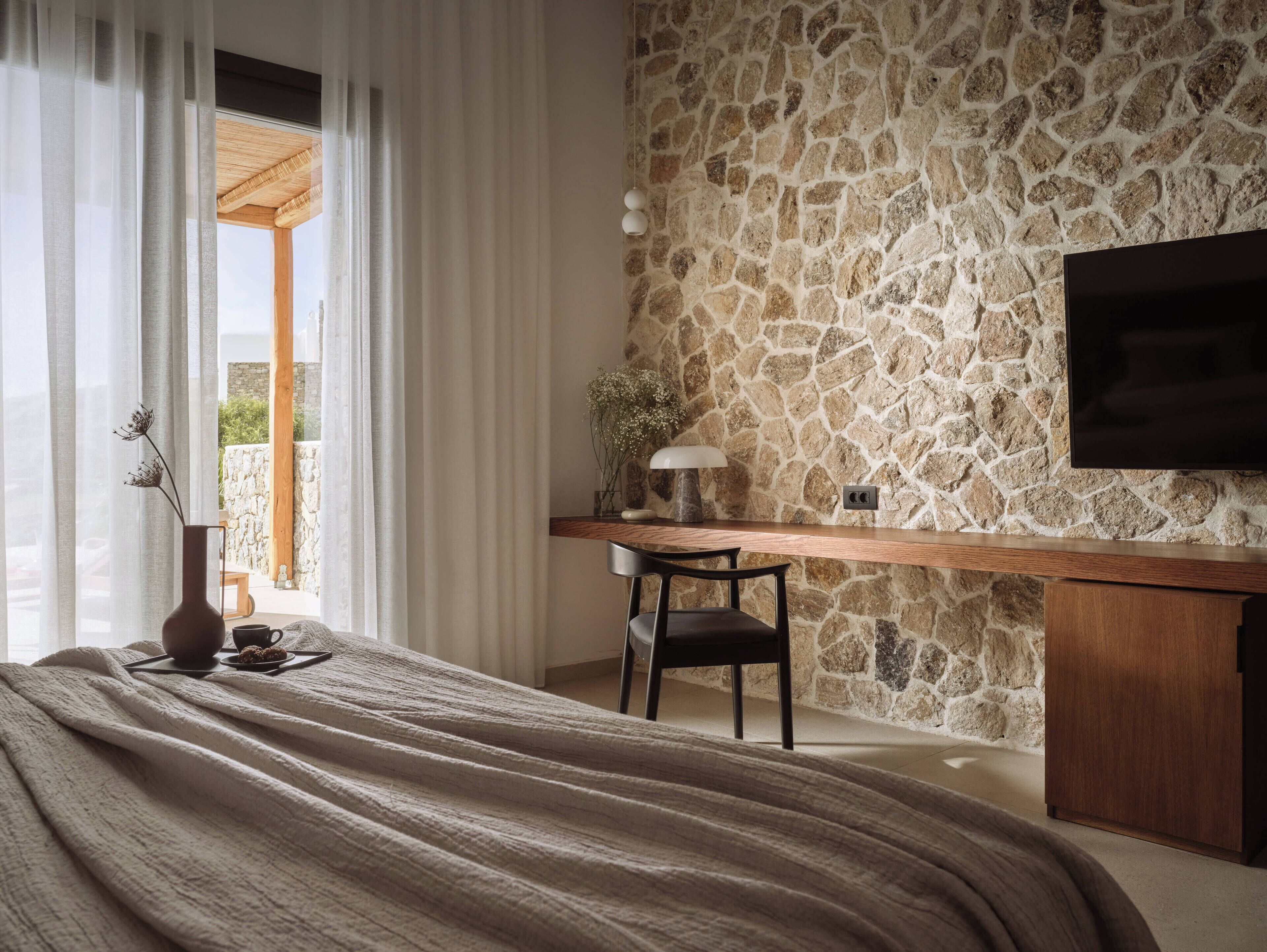 Un dormitorio con pared de piedra, un escritorio de madera, una silla, un televisor montado en la pared y una cama con manta.