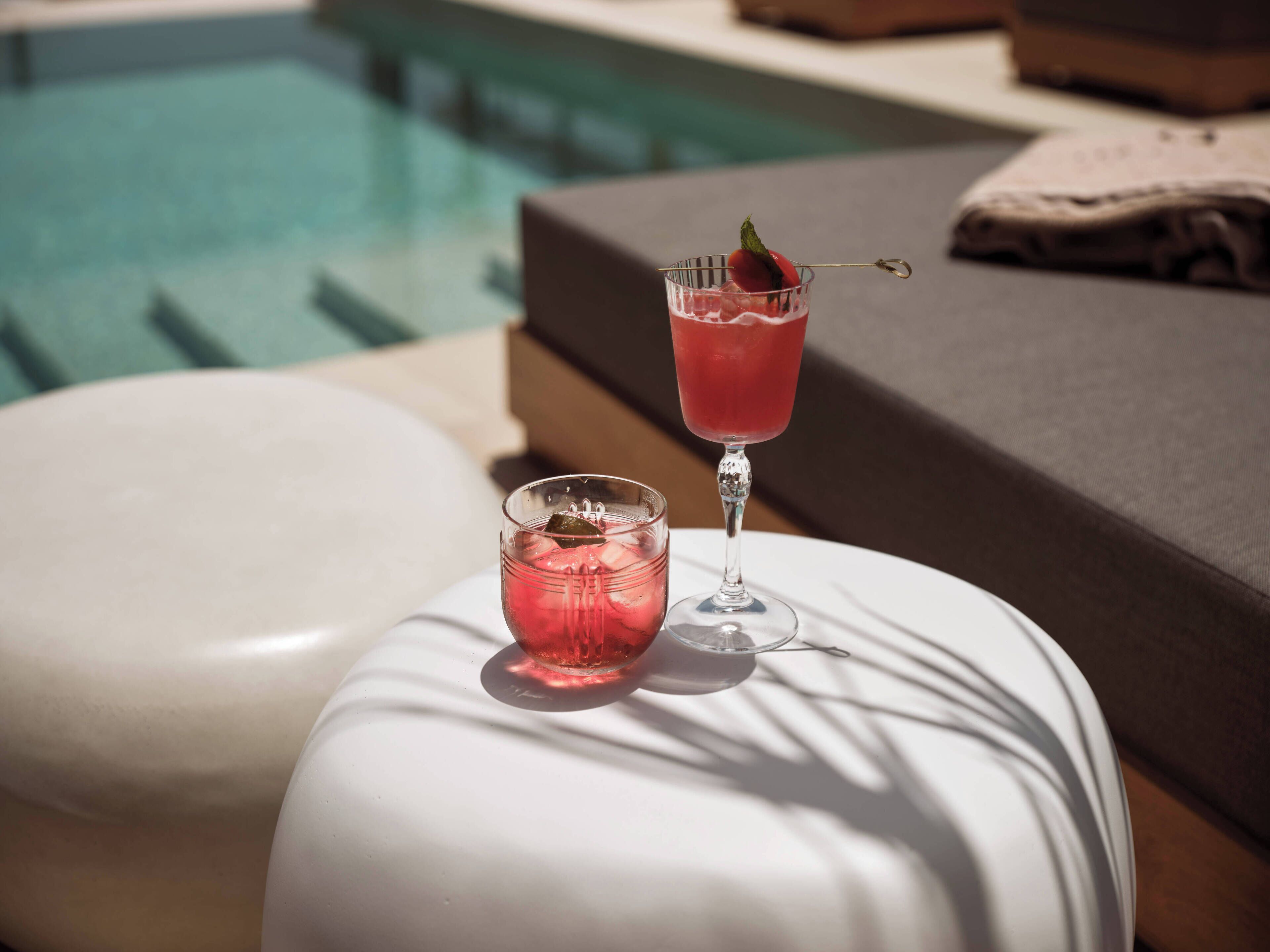 Une scène de bord de piscine avec un verre de cocktail rouge et un verre à cocktail rempli de glaçons.