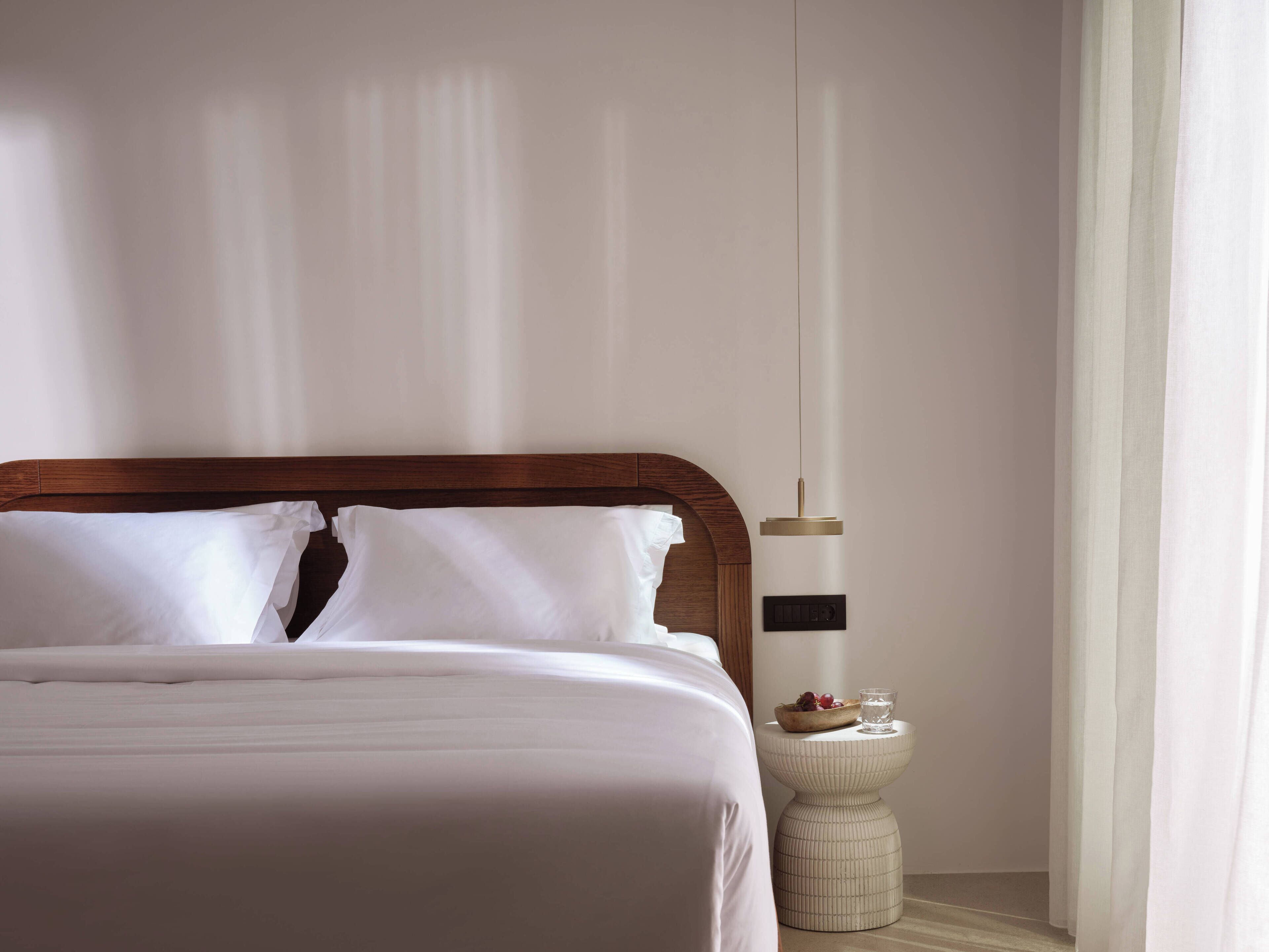 A neatly made bed with white linens, a wooden headboard, a bedside table with a bowl of fruit and a glass, and a wall-mounted light fixture.