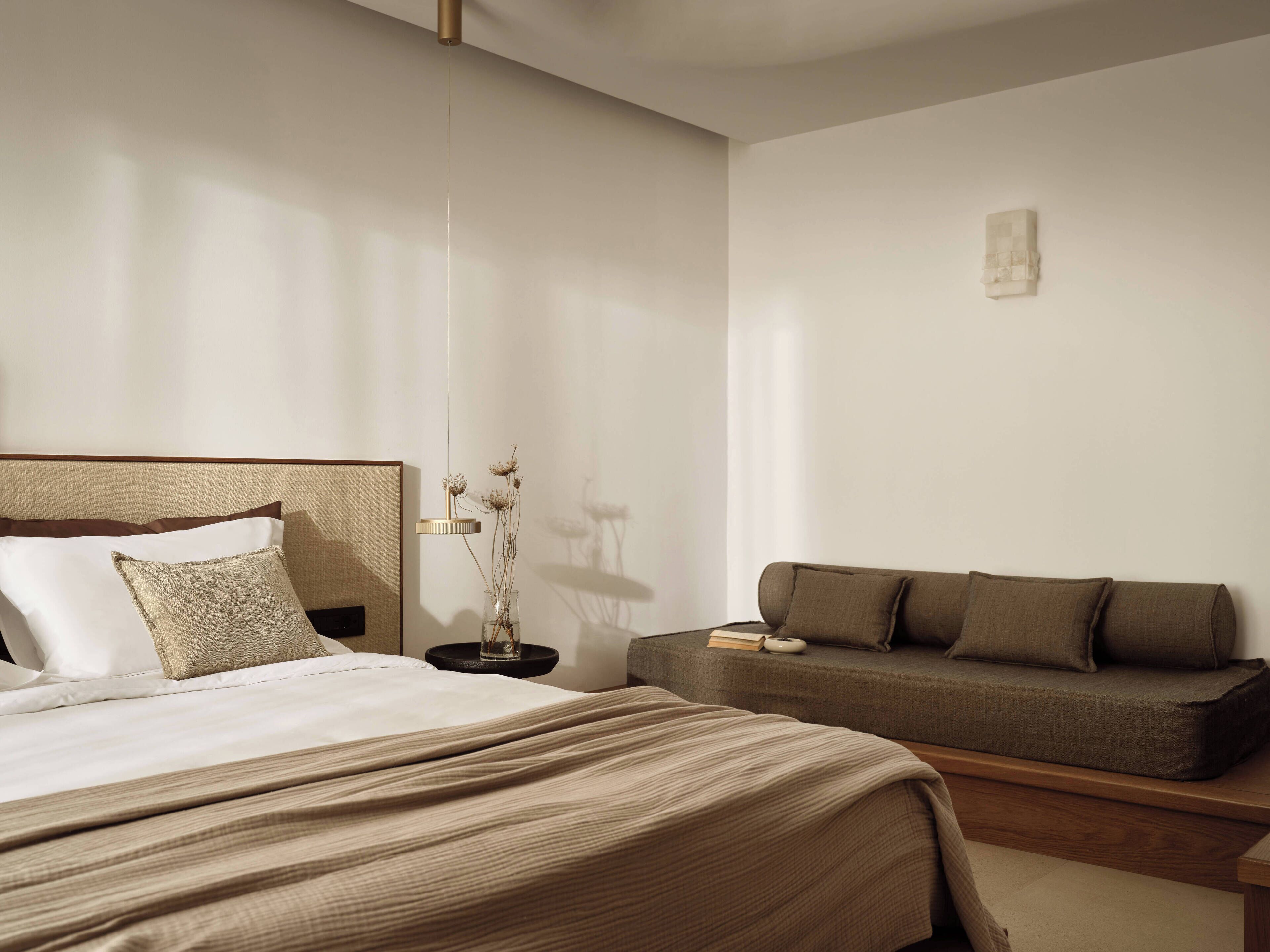 A modern hotel room with a large bed, a sofa, a bedside table with a vase of dried flowers, and a small artwork on the wall.