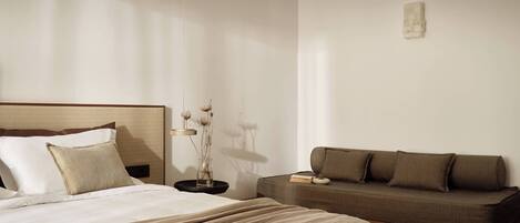 A modern hotel room with a large bed, a sofa, a bedside table with a vase of dried flowers, and a small artwork on the wall.