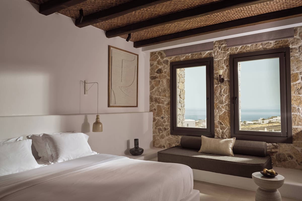 A bedroom with a bed, a stone wall, a window with a view, and a wall-mounted lamp.