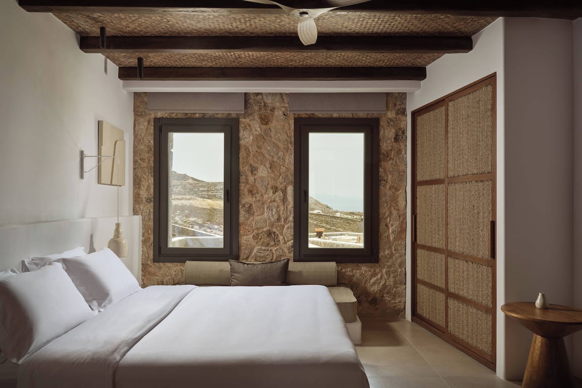 A bedroom with a large bed, two windows with a view of a mountainous landscape, a wooden ceiling, and a traditional woven partition.