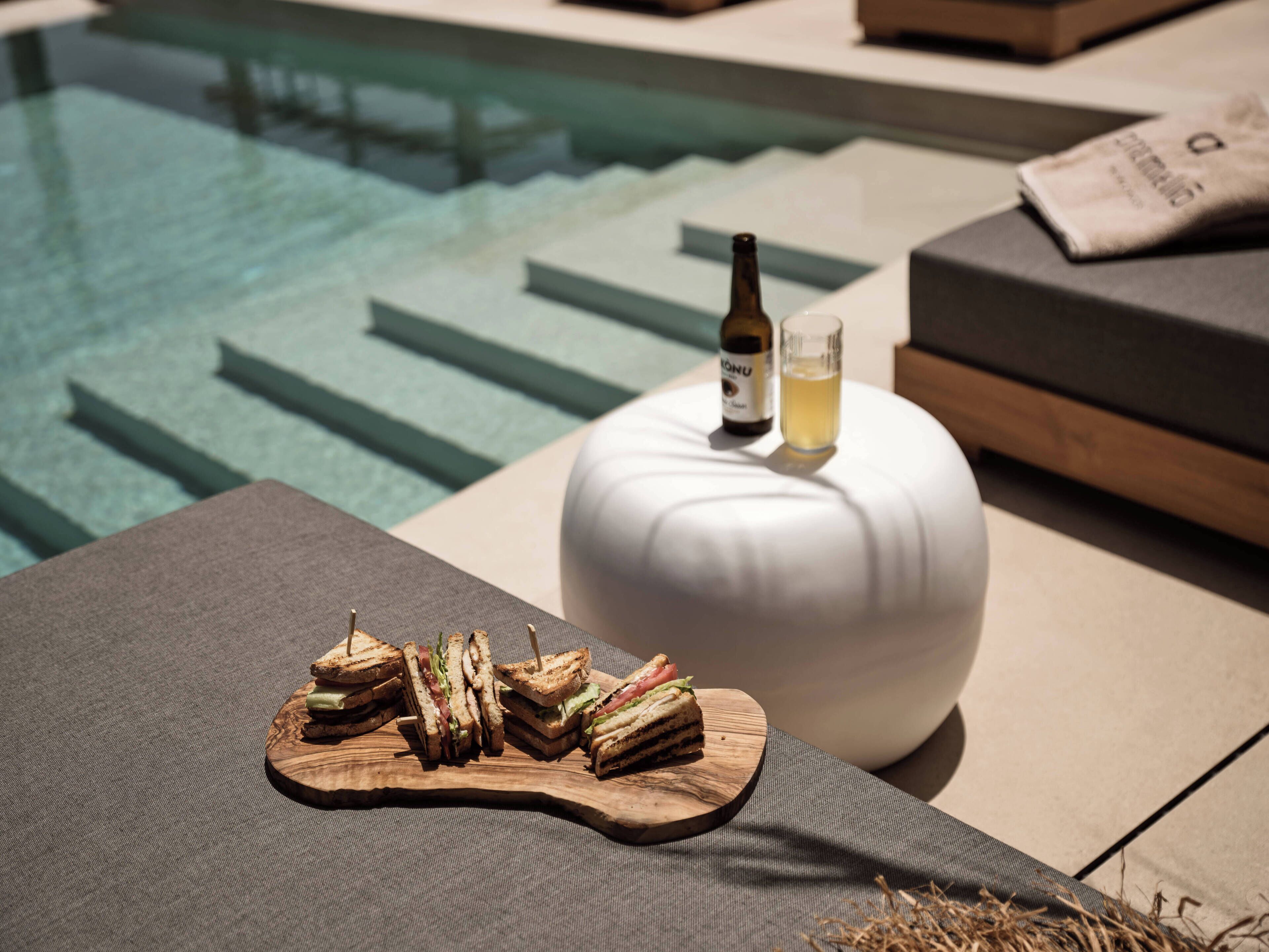 A poolside setting with a wooden board holding sandwiches, a bottle of drink, and a glass of water on a white round table.