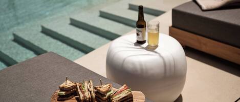 A poolside setting with a wooden board holding sandwiches, a bottle of drink, and a glass of water on a white round table.