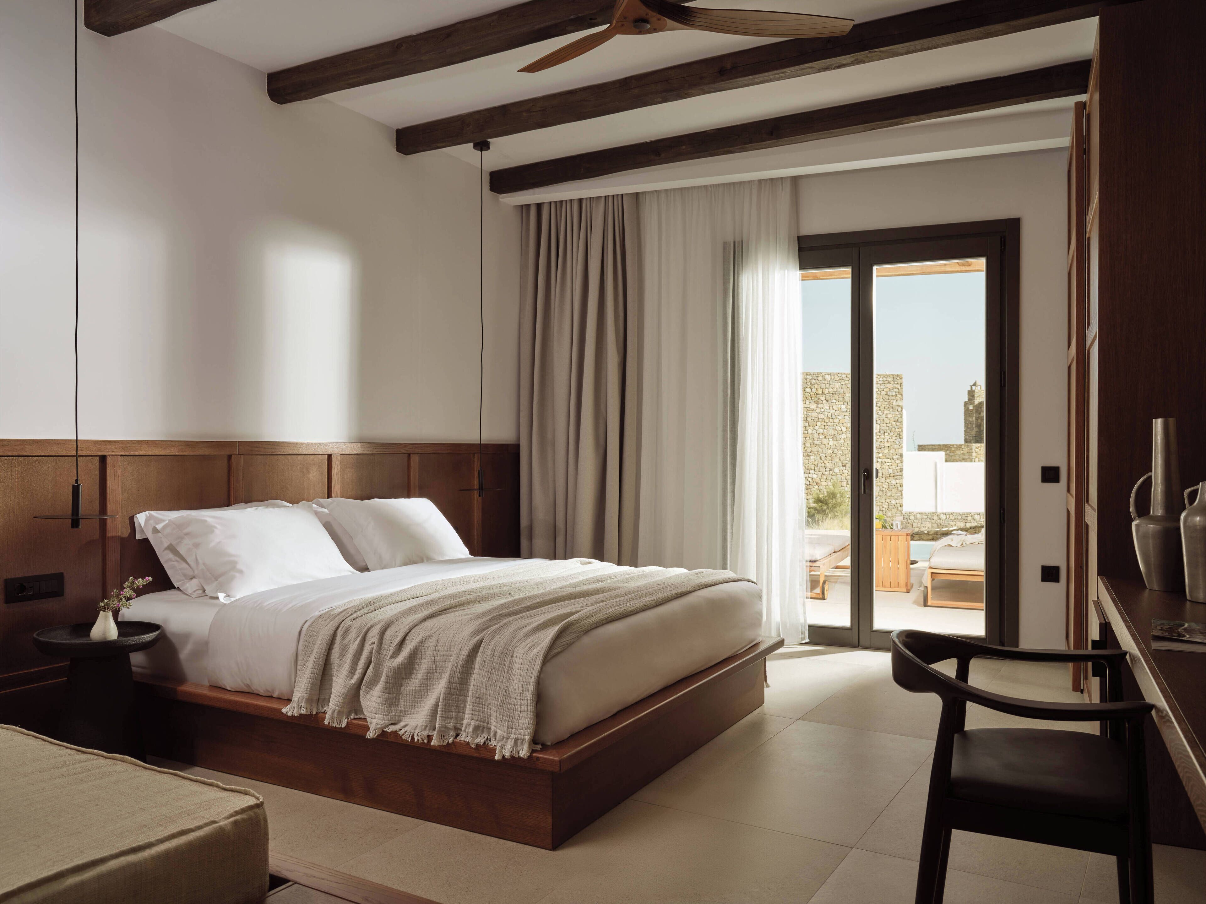 A bedroom with a wooden headboard, a bed with white linens, a bedside table with a vase, and a view of an outdoor balcony.