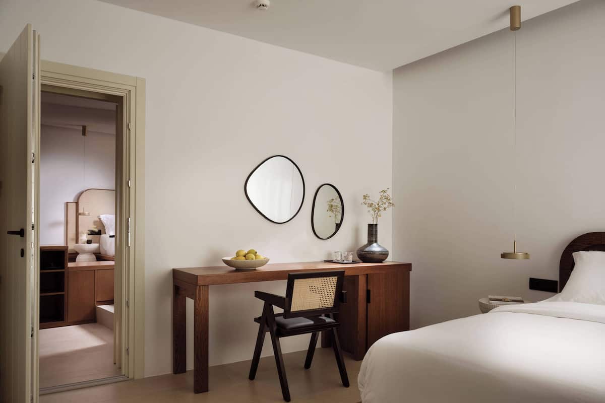 A bedroom with a bed, a desk with a chair, a bedside table, a mirror, and a vase with flowers.