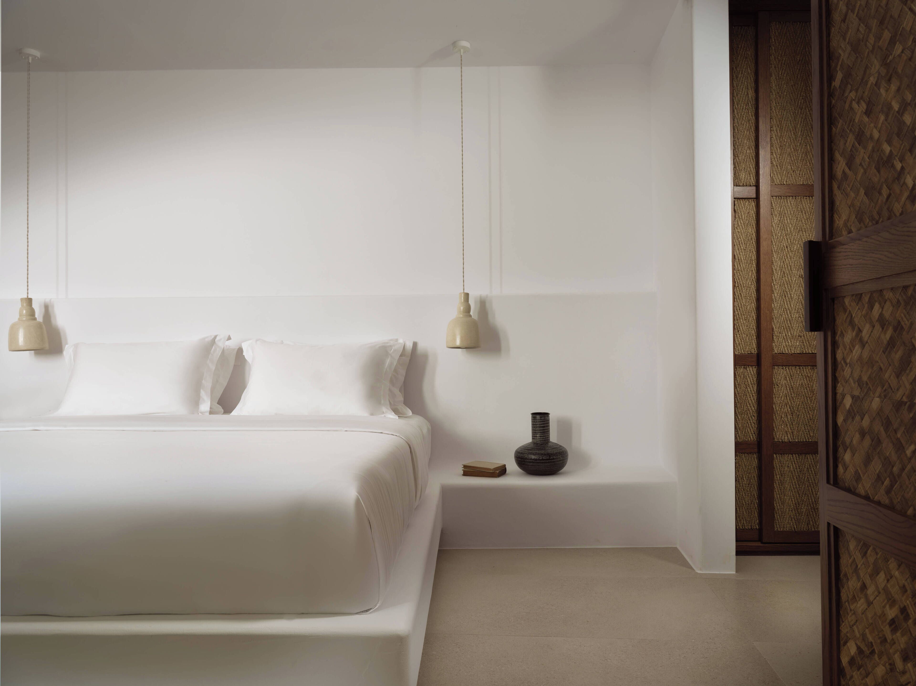 A modern bedroom with a large bed, two bedside lamps, a small table with a vase, and a woven partition.