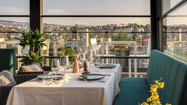 A dining area with a table set for two, a potted plant, and a view of the cityscape through large windows. - Vibre Hotel (Cluj-Napoca)