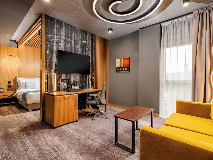A modern hotel room with a bed, a desk with a flat-screen TV, a coffee machine, and a yellow armchair.
