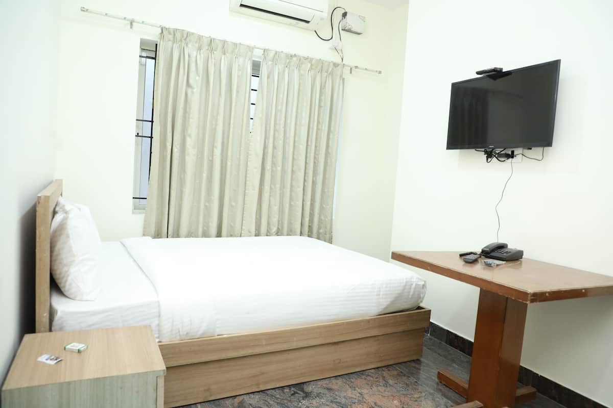A bedroom with a bed, a wooden nightstand, a desk, a television, and a window with curtains.