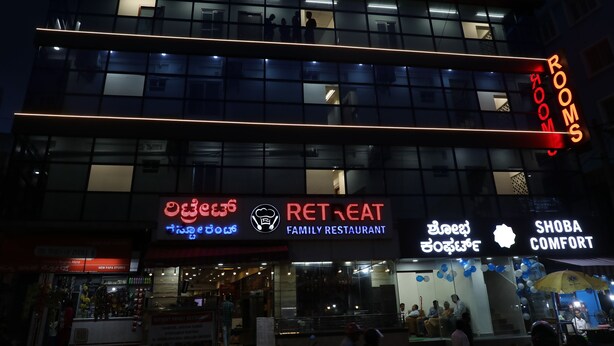 A multi-story building with a 'Retreat Family Restaurant' sign, 'Rooms' sign, and 'Shoba Comfort' sign. - Shoba Comfort (Bengaluru)