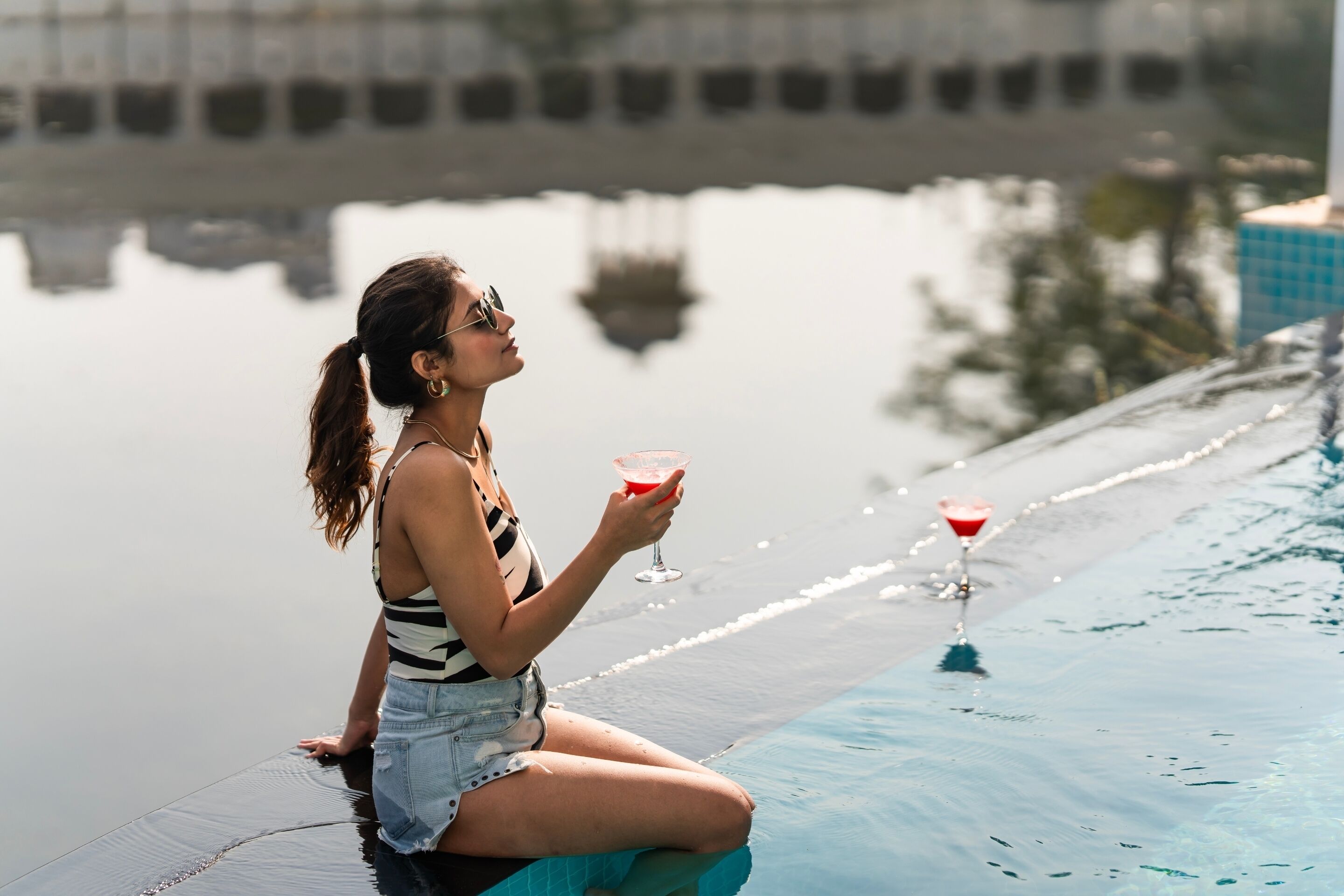 Une personne assise au bord d’une piscine, un verre à la main.