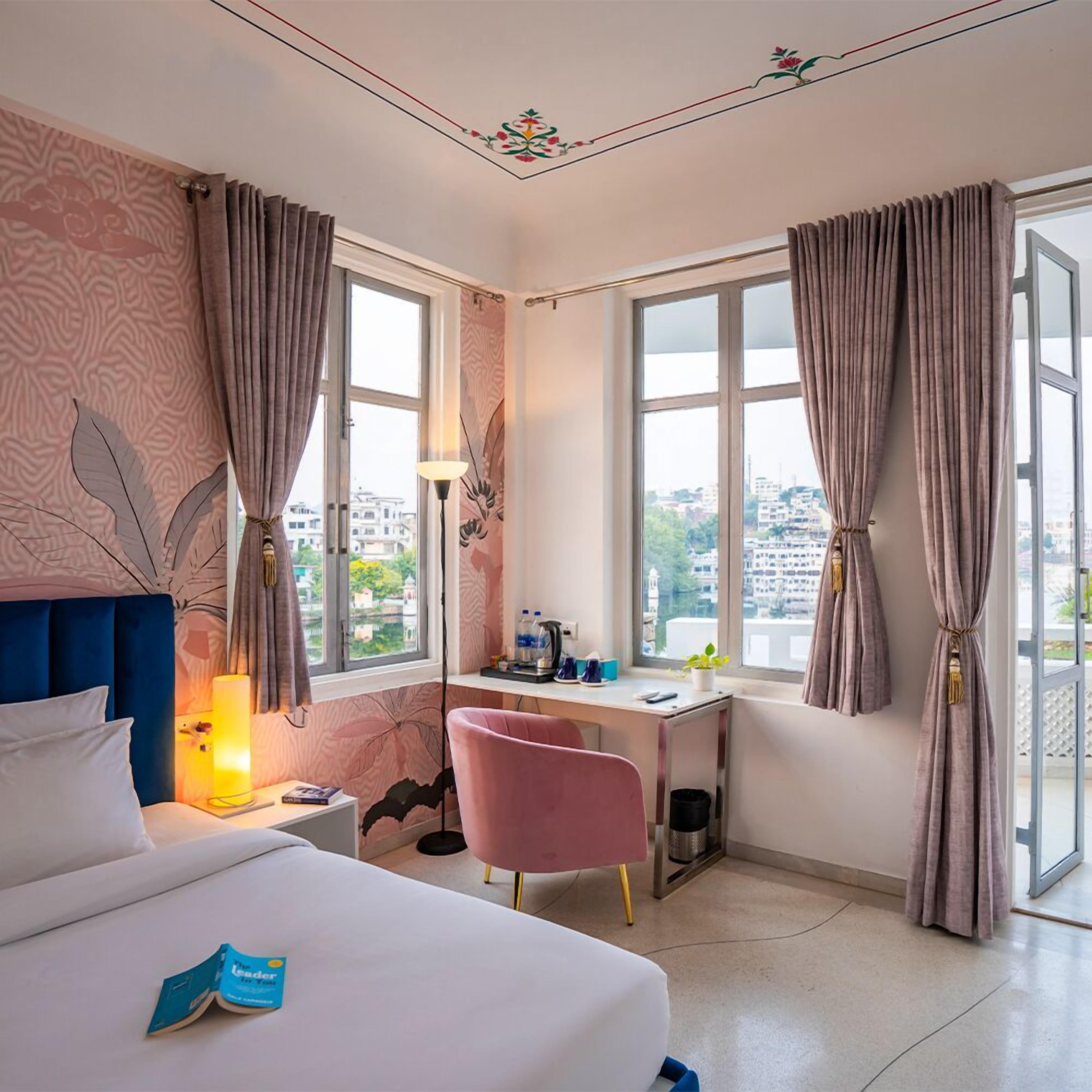Hotelkamer met een bed, een bureau, een stoel, een lamp en uitzicht op de stad door grote ramen.