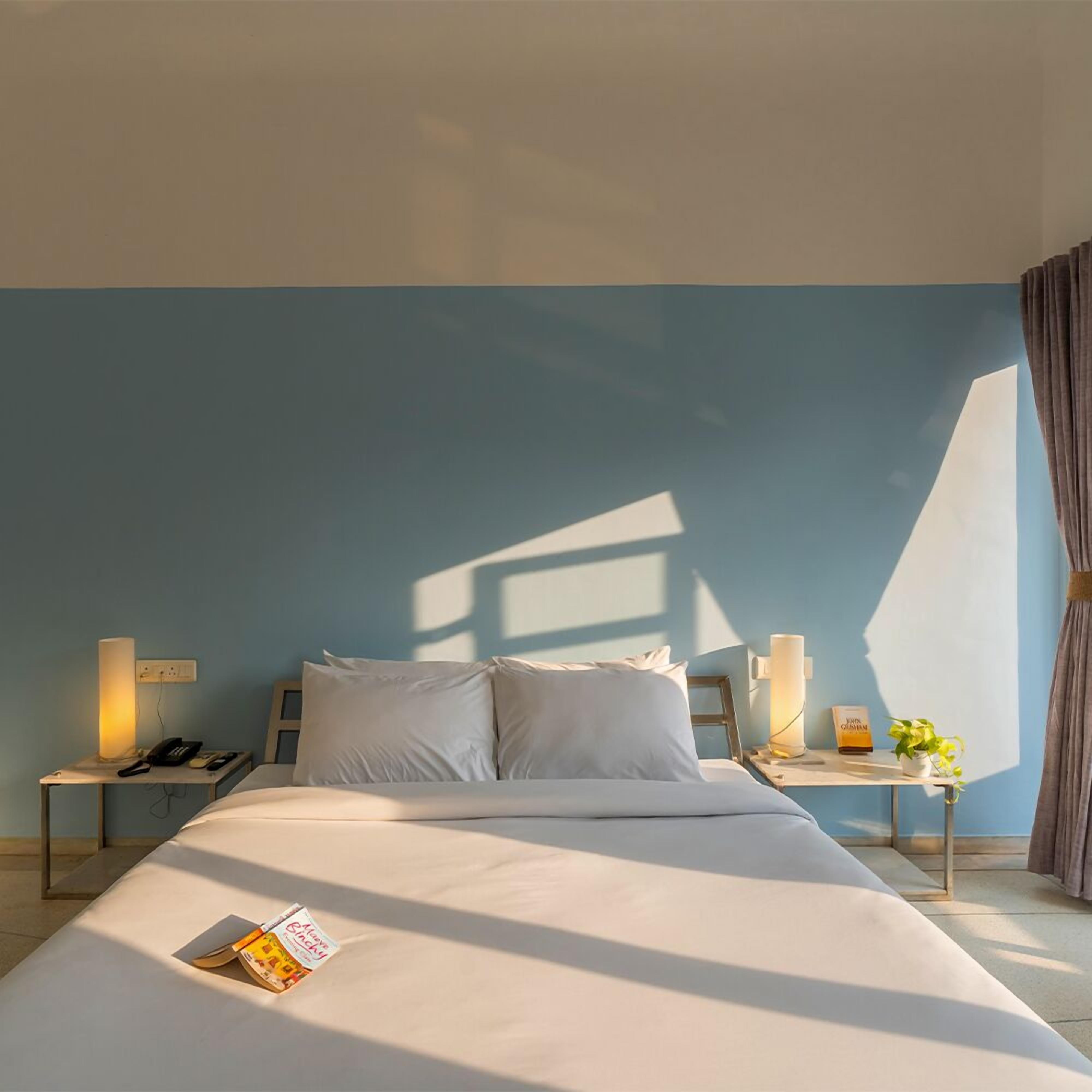 Ein ordentlich gemachtes Bett in einem Zimmer mit blauer Wand, zwei Nachttischen inklusive Lampen und ein Fenster, das einen Schatten wirft.
