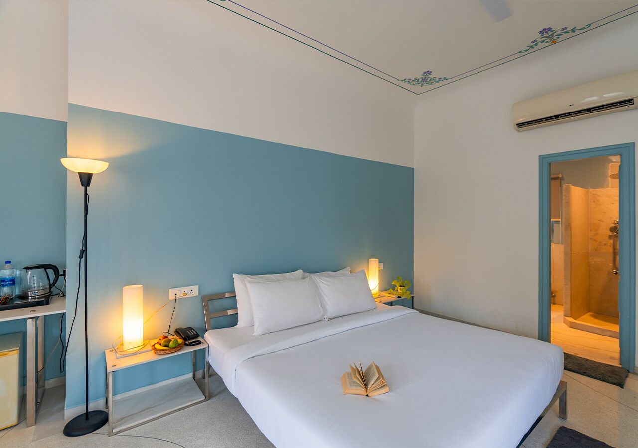 A hotel room with a bed, bedside lamps, a desk with a lamp, and an air conditioning unit.