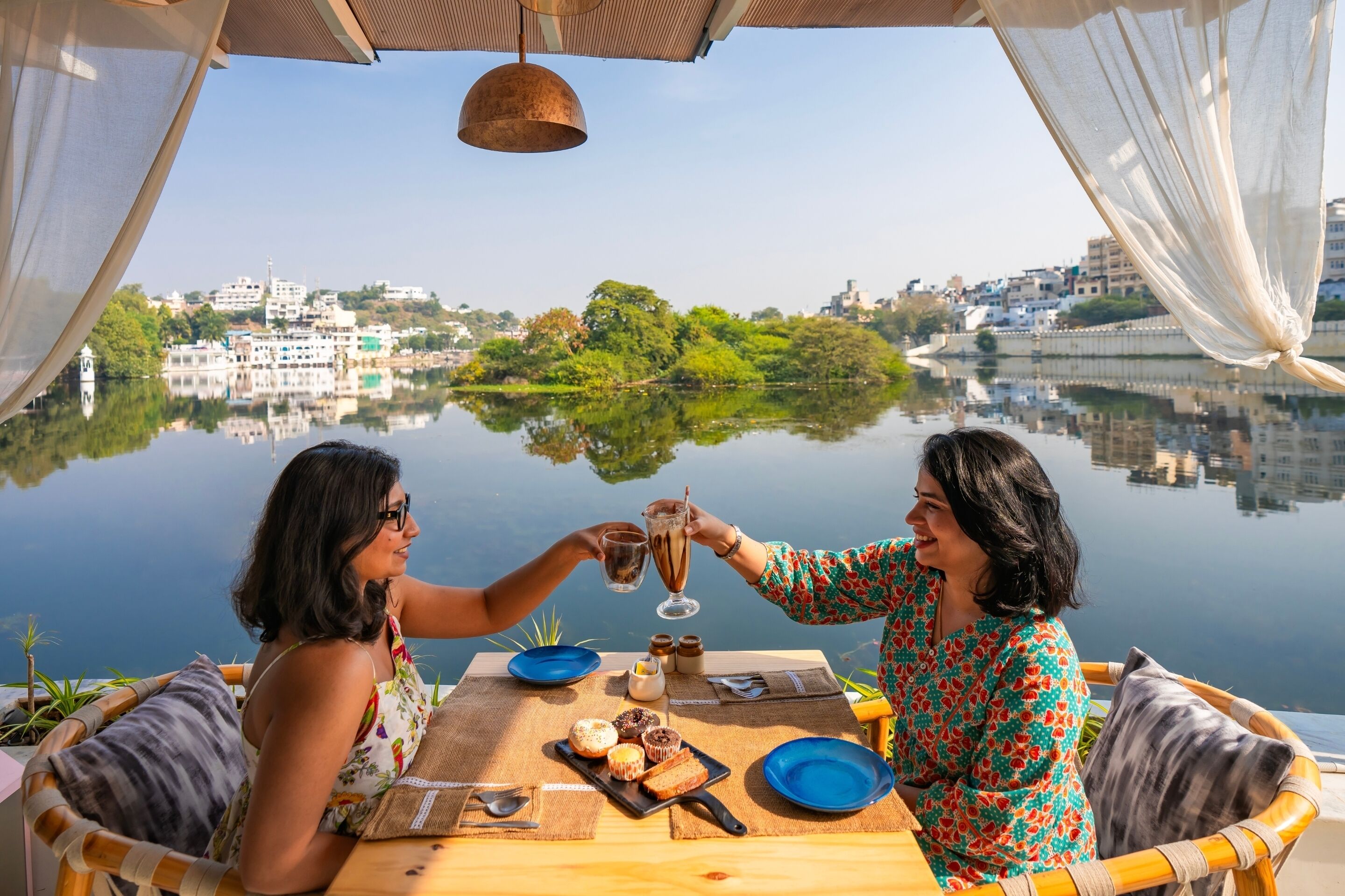 Deux personnes trinquent à une table, avec une vue sur un lac et des bâtiments en arrière-plan.