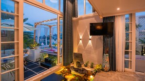 A modern living room with large windows, a flat-screen TV, a stone fireplace, and a small indoor garden. - Moustache Udaipur Verandah (Udaipur)