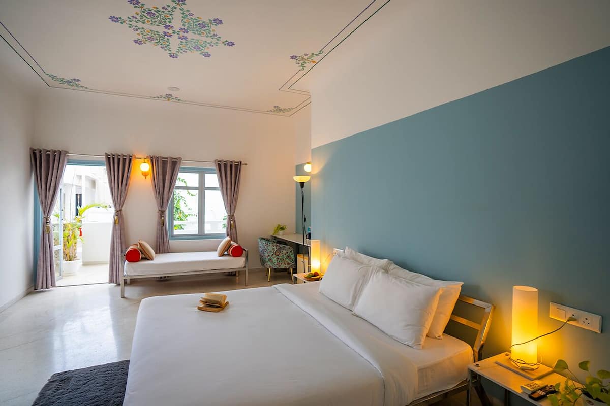 A hotel room with a large bed, a bedside table with a lamp, a window with curtains, and a ceiling with floral decorations.