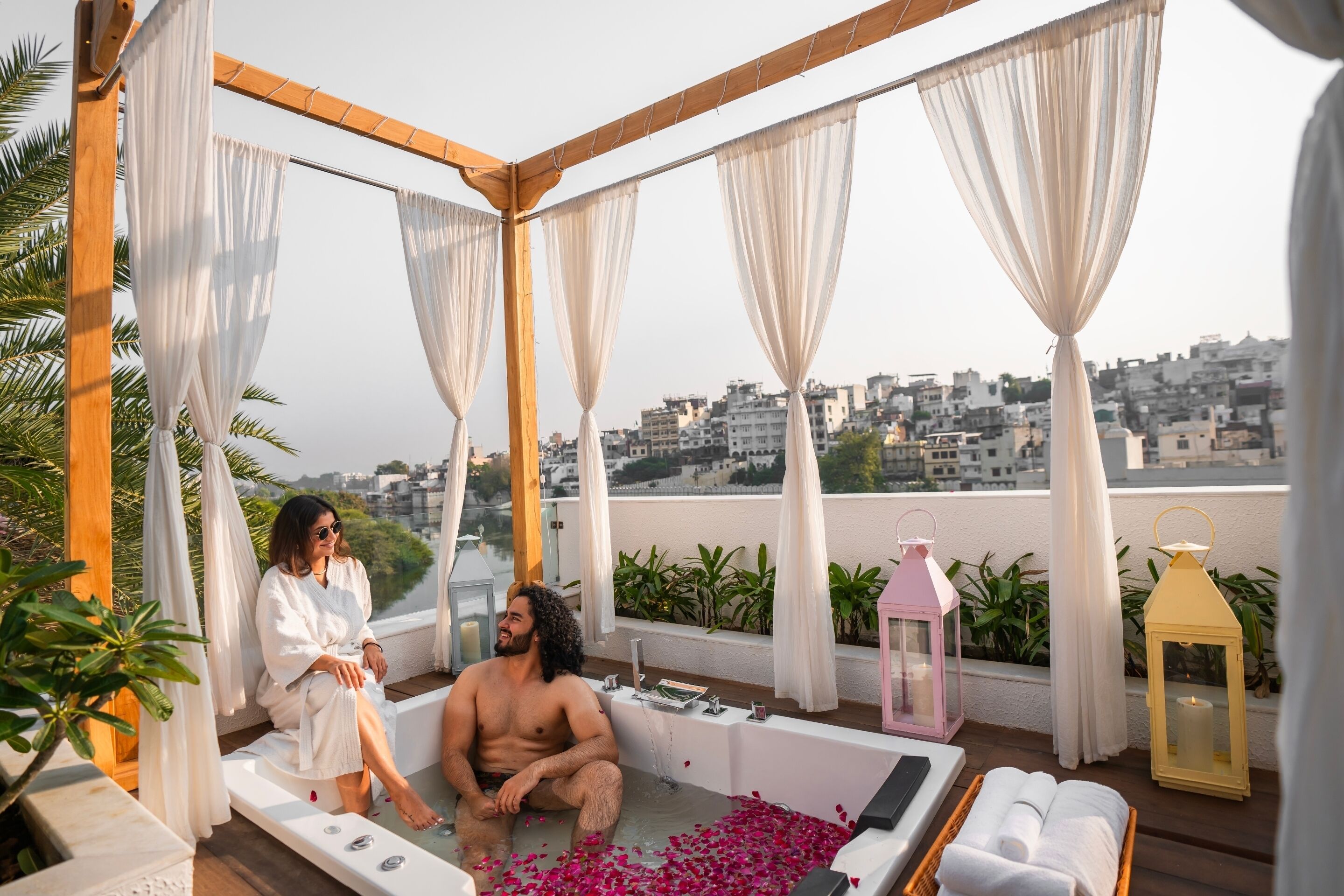 A rooftop terrace with a bathtub, white curtains, and two people relaxing.