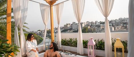 Terraço no telhado com banheira, cortinas brancas e duas pessoas relaxando.