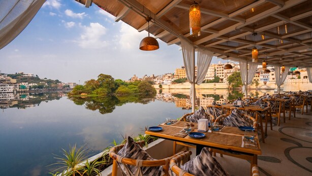 An outdoor dining area with tables and chairs, overlooking a calm body of water and a cityscape. - Moustache Udaipur Verandah (Udaipur)