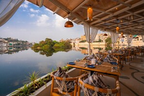 Un área de comedor al aire libre con mesas y sillas, con vista a un cuerpo de agua tranquilo y un paisaje urbano.