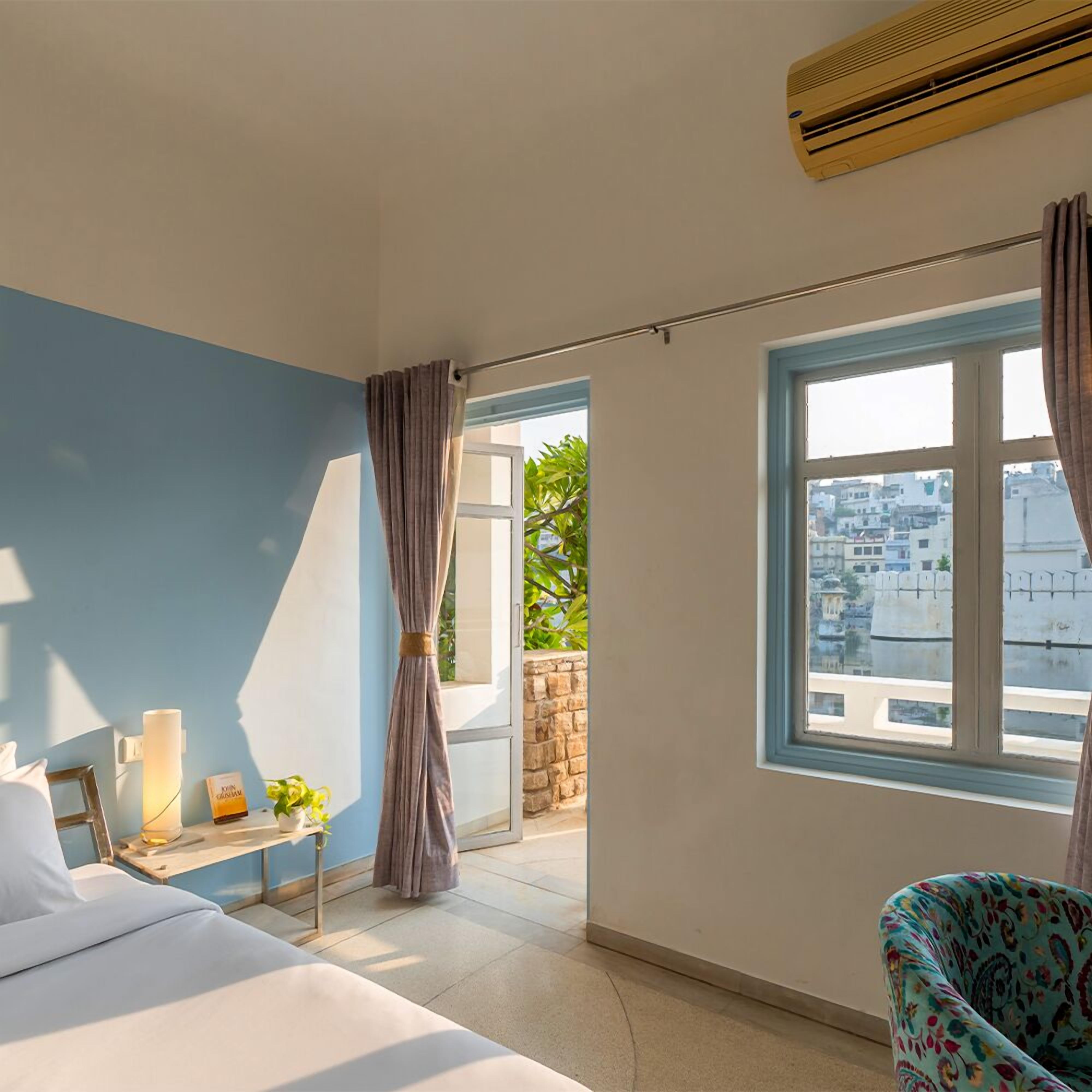 A bedroom with a bed, a bedside table with a lamp, a window with curtains, and a view of the cityscape.