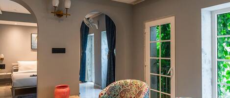 A modern living room with a sofa, a small table with a bowl of fruit, a mirror, and a window with a view of greenery.