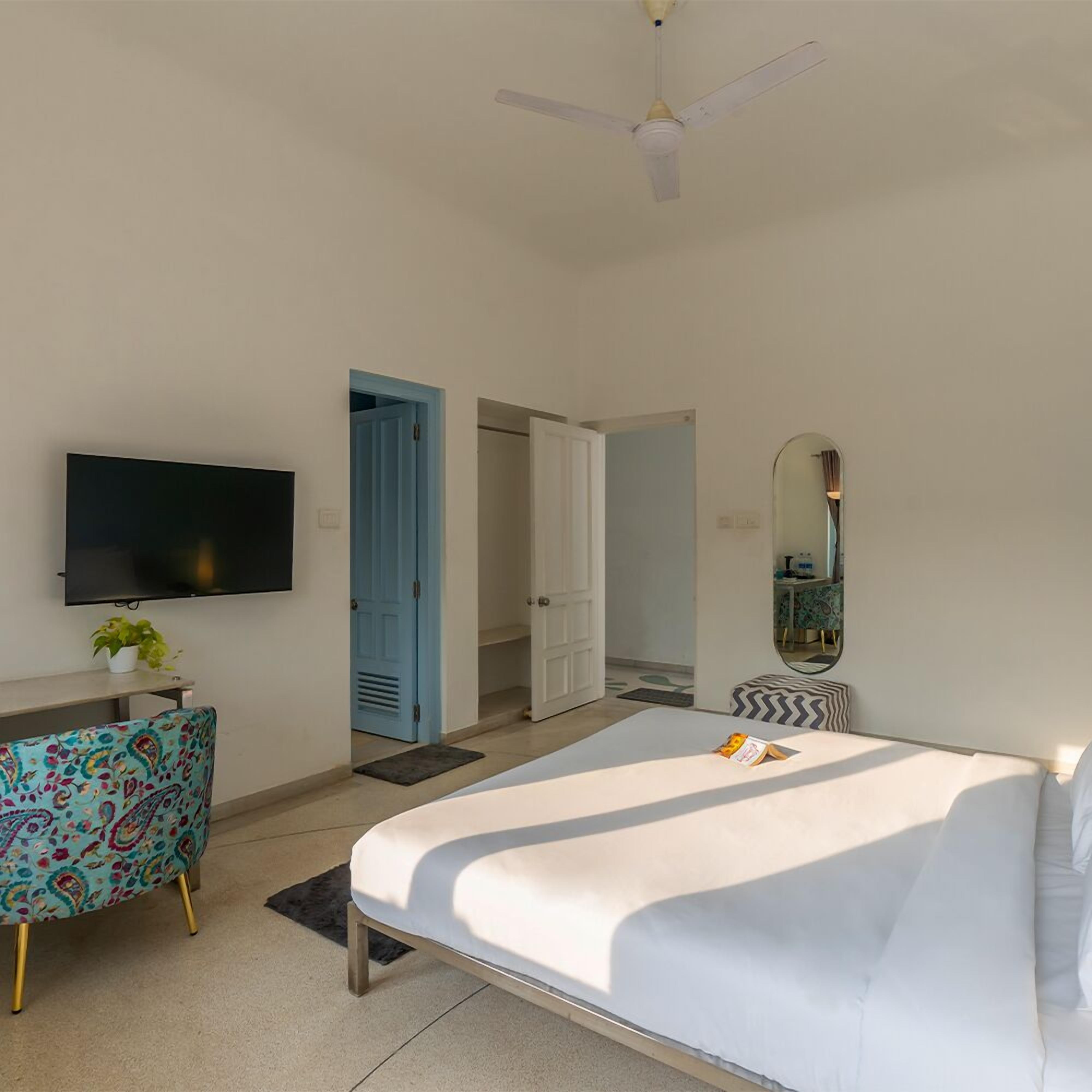 A hotel room with a bed, a desk with a TV, a chair, a ceiling fan, and a door leading to another room.