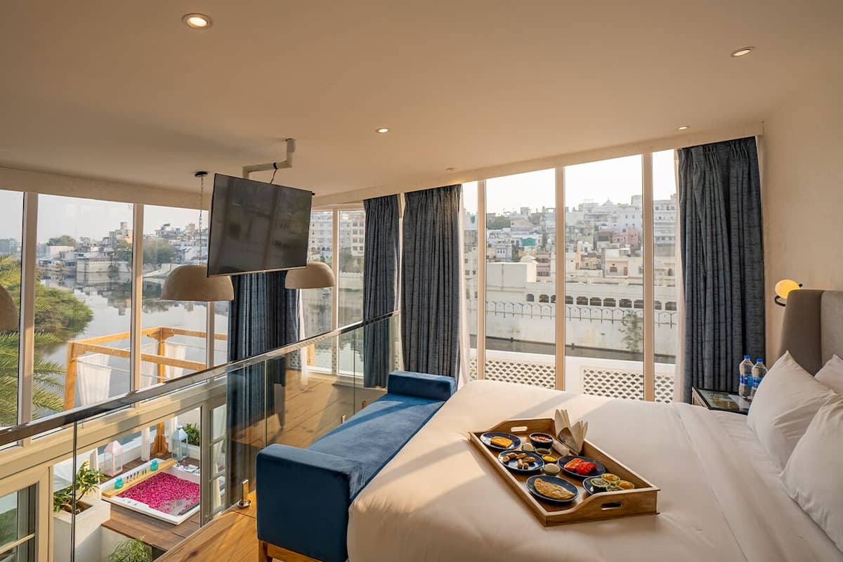 A hotel room with a large bed, a balcony with a view of the city, and a tray of breakfast items on the bed.