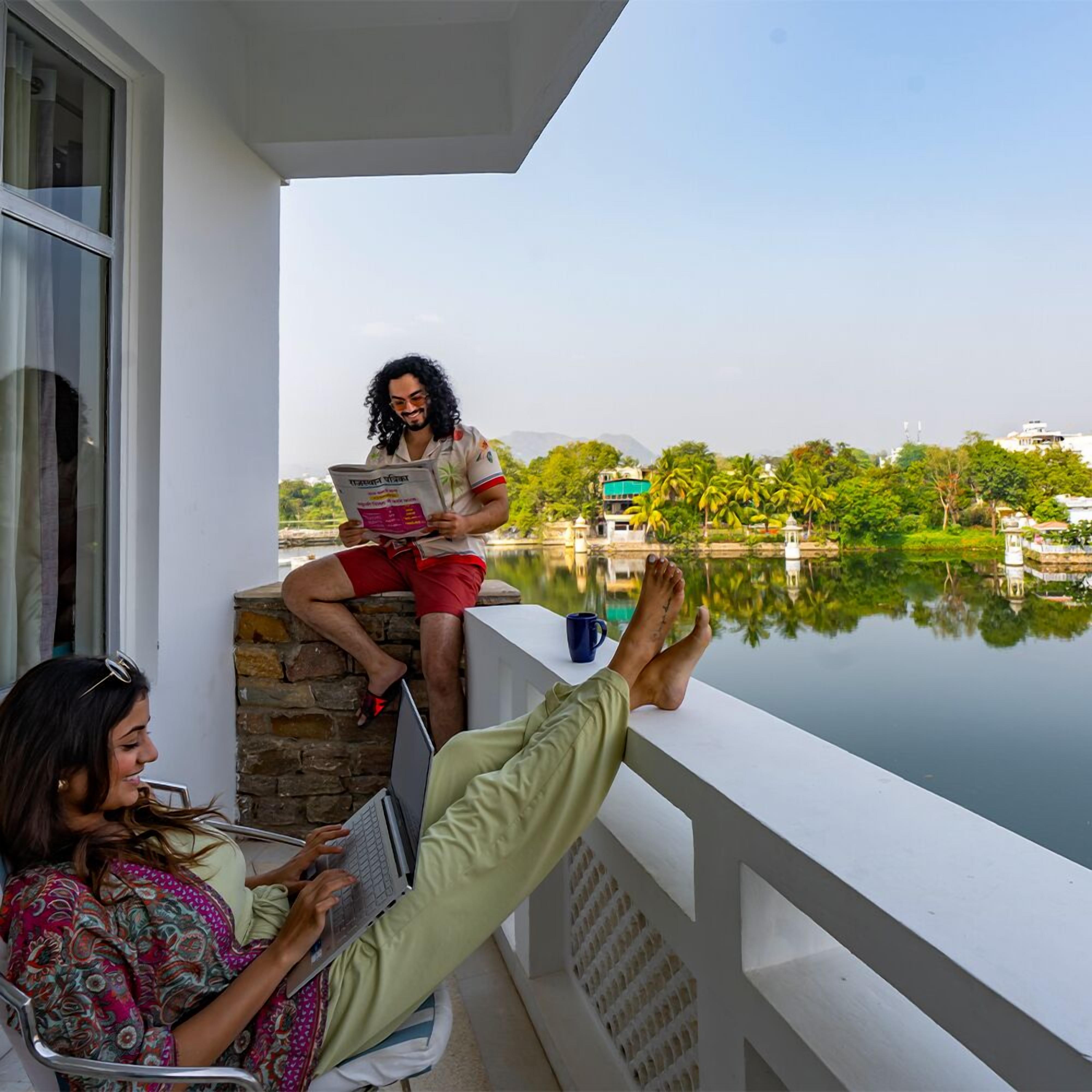Un balcone con vista su un lago e degli edifici, una persona legge un giornale e un'altra utilizza un portatile.