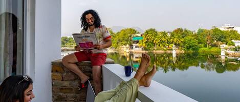 Un balcone con vista su un lago e degli edifici, una persona legge un giornale e un'altra utilizza un portatile.