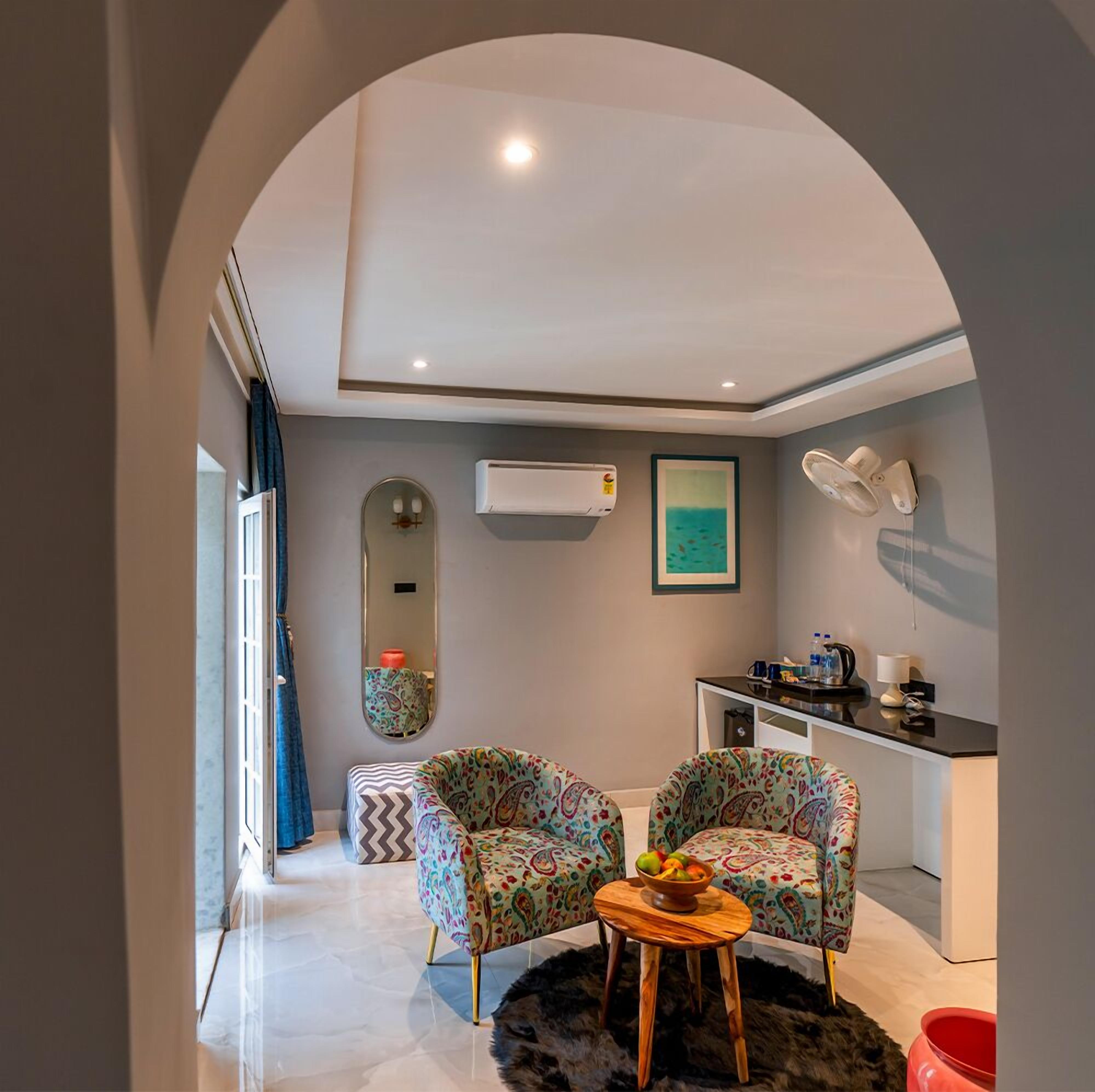 A compact living space with two patterned armchairs, a small wooden table, a mirror, an air conditioning unit, a painting, and a kitchenette.