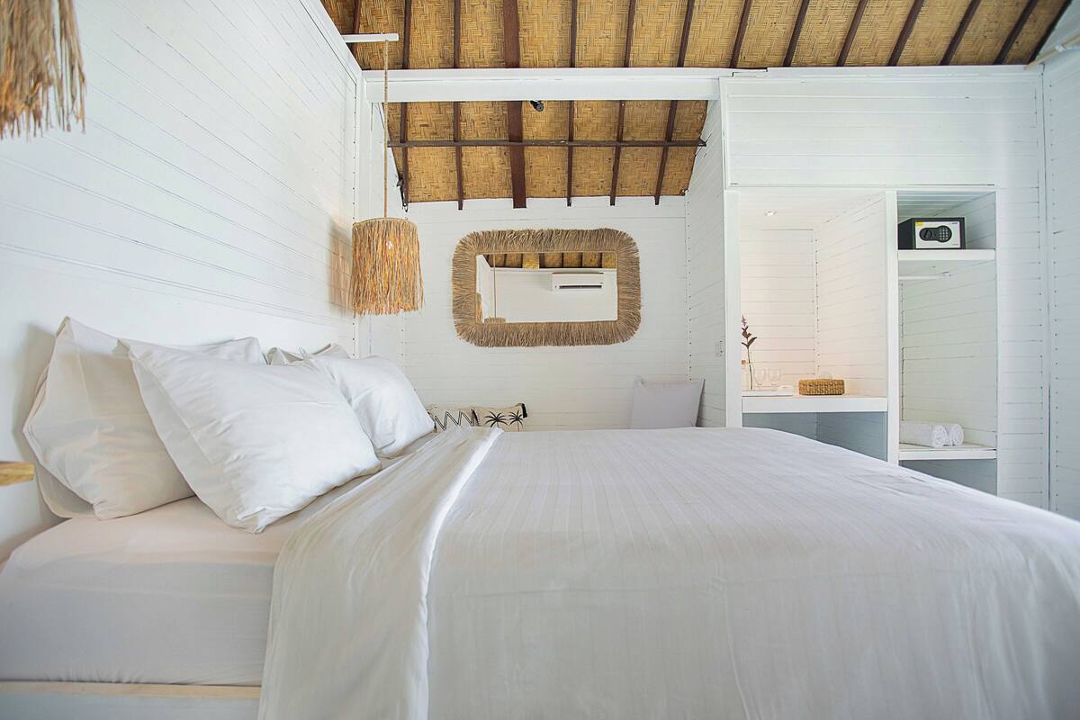 A bedroom with a bed, white walls, a wooden ceiling, a mirror, and a shelf with toiletries.