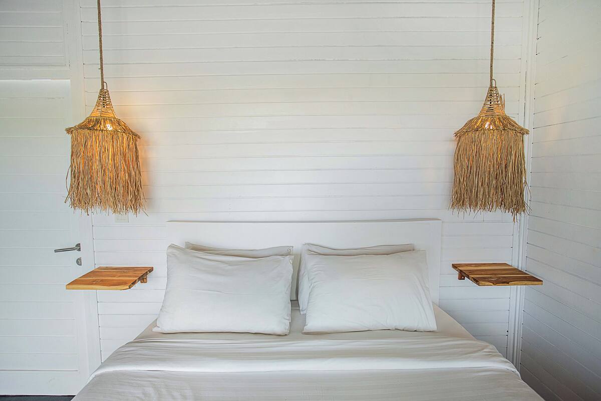 A bed with white bedding, two bedside tables, and two hanging lamps with a woven design.