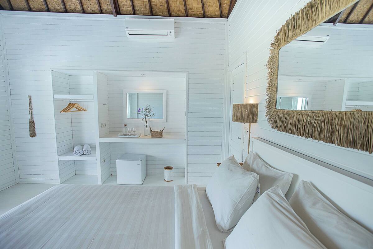 A white room with a bed, a mirror, a shelf with towels, and a small table with a vase.