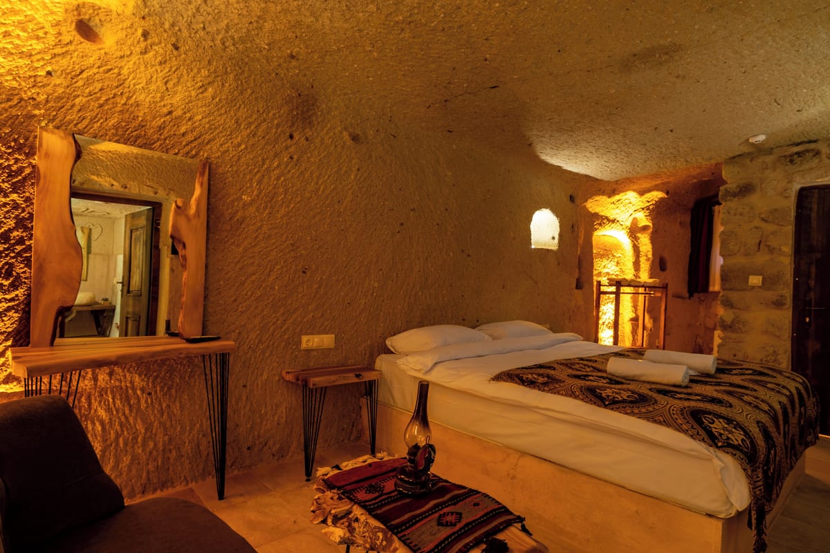 A rustic room with a bed, a mirror, a small table, and a wall-mounted light fixture.
