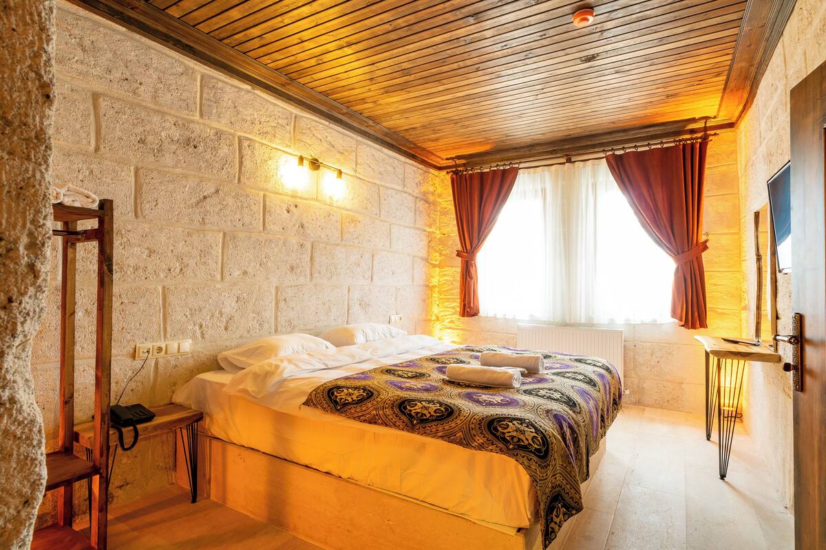 A bedroom with a wooden ceiling, a brick wall, a bed with patterned bedding, a bedside table, and a window with curtains.