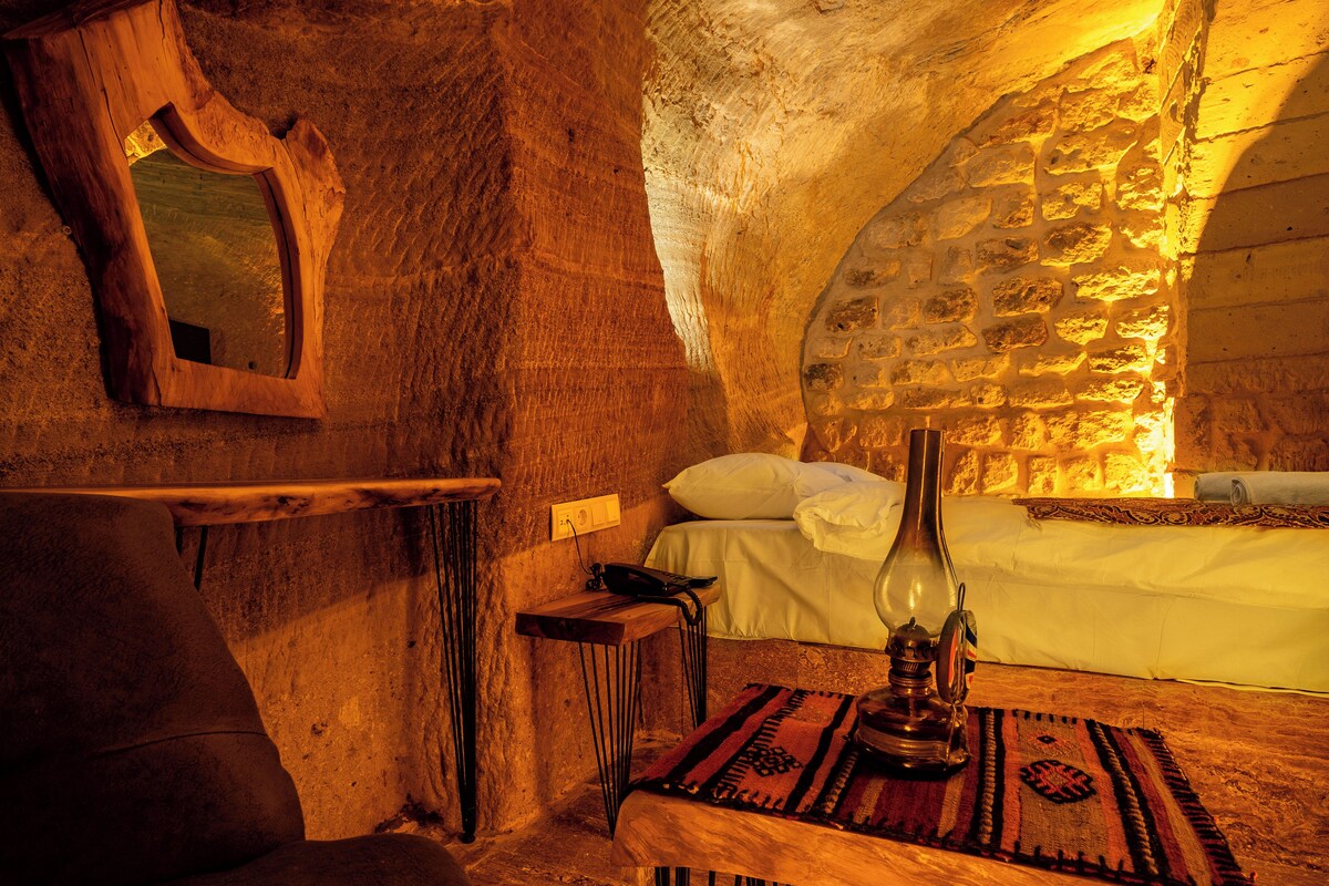 A rustic stone interior with a wooden bed, a small table with a lamp, and a mirror on the wall.