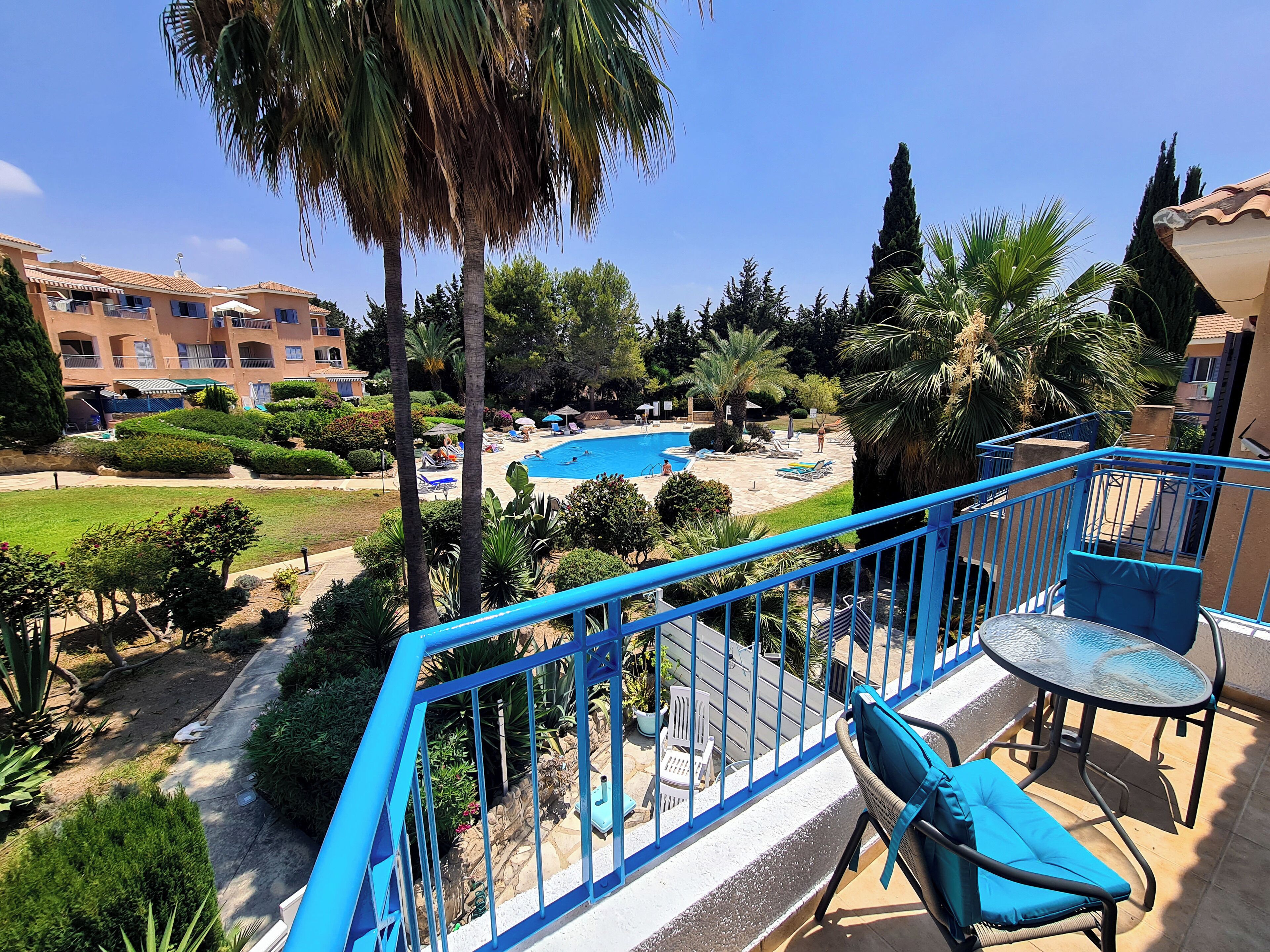 A balcony with blue railings overlooking a swimming pool, surrounded by palm trees and a landscaped garden.