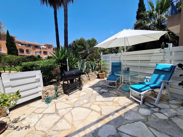 An outdoor patio with stone flooring, a barbecue grill, two blue lounge chairs, a white umbrella, and a white fence.