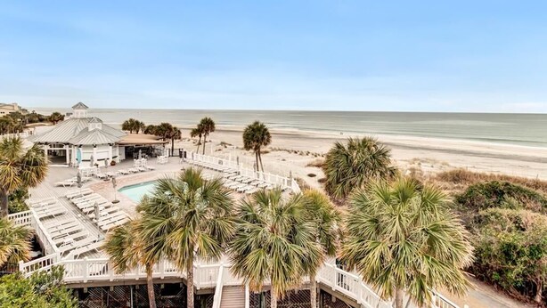 A beachfront hotel with a pool, lounge chairs, and palm trees. - Sandy Toes | Iconic Oceanfront Home with Sunset Views at Grand Pavilion (Isle of Palms)