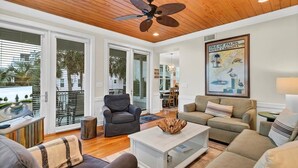 A spacious living room with a wooden ceiling, large windows, a ceiling fan, and a framed map of the Isle of Palms. - Sandy Toes | Iconic Oceanfront Home with Sunset Views at Grand Pavilion (Isle of Palms)