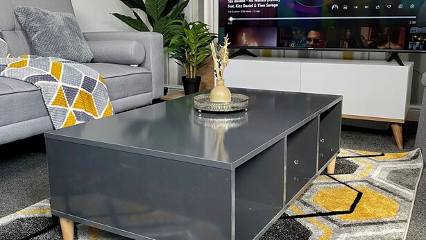 A modern living room with a grey sofa, a coffee table, and a TV displaying a music video. - Gillingham cosy 3 bedroom with free WiFi & parking (Gillingham)