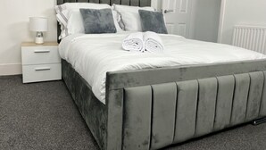 A neatly made bed with grey headboard and footboard, white bedding, and two grey pillows. There is a bedside table with a lamp and a drawer. - Gillingham cosy 3 bedroom with free WiFi & parking (Gillingham)