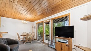 A room with a wooden ceiling, a flat-screen TV on a wooden stand, a dining table with chairs, and a sofa.