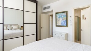 A bedroom with a bed, a dresser, a painting, and a door leading to another room. - MAMAD Airy & Bright with Balcony & Parking steps from Beach by FeelHome (Tel Aviv - Jaffa)